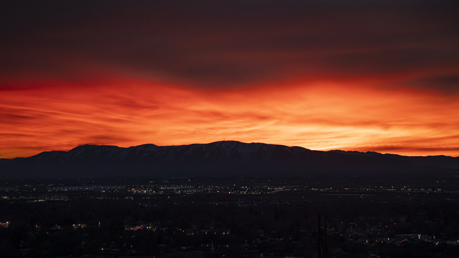 The clouds glow brilliant yellow and red above West Mountain in silhouette with Provo town lights below